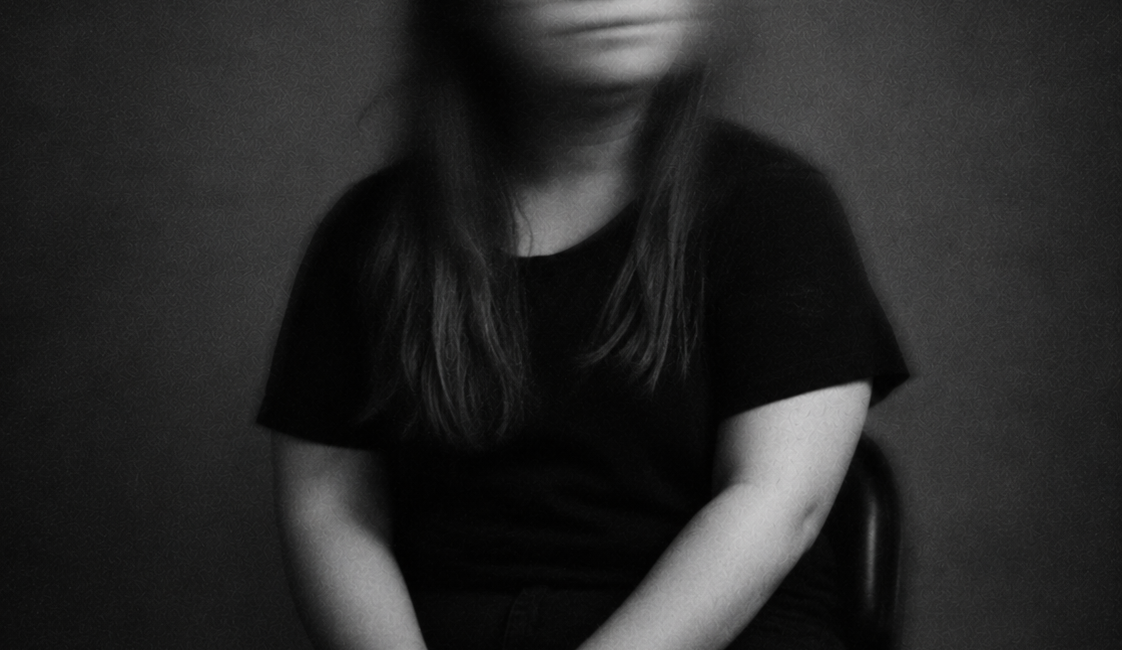 Sitzende Person mit langen dunklen Haaren und dunkler Kleidung, die die Hände im Schoß hält. Das nach vorne gerichtete Gesicht ist durch Bewegungsunschärfe verschwommen.