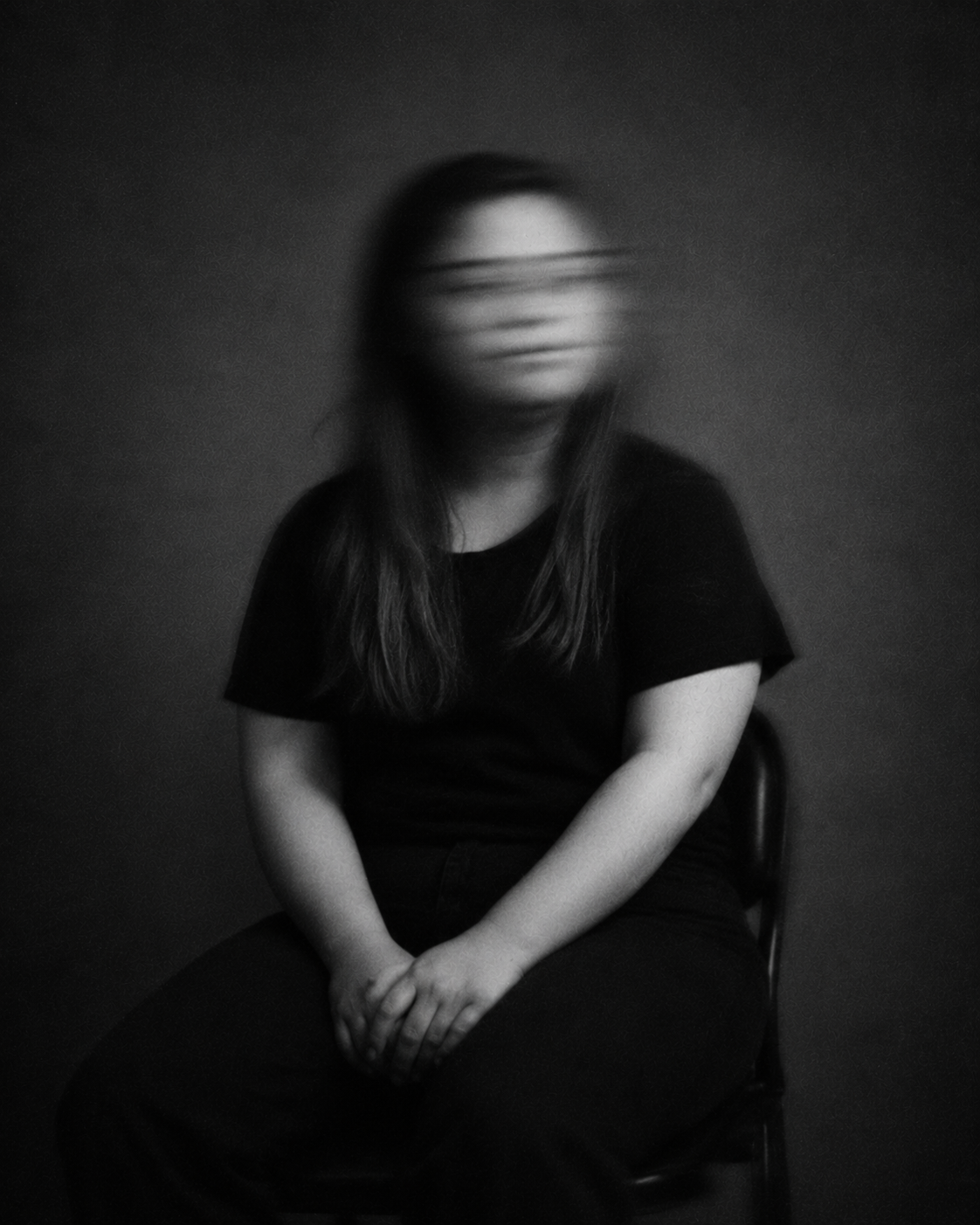 Sitzende Person mit langen dunklen Haaren und dunkler Kleidung, die die Hände im Schoß hält. Das nach vorne gerichtete Gesicht ist durch Bewegungsunschärfe verschwommen.