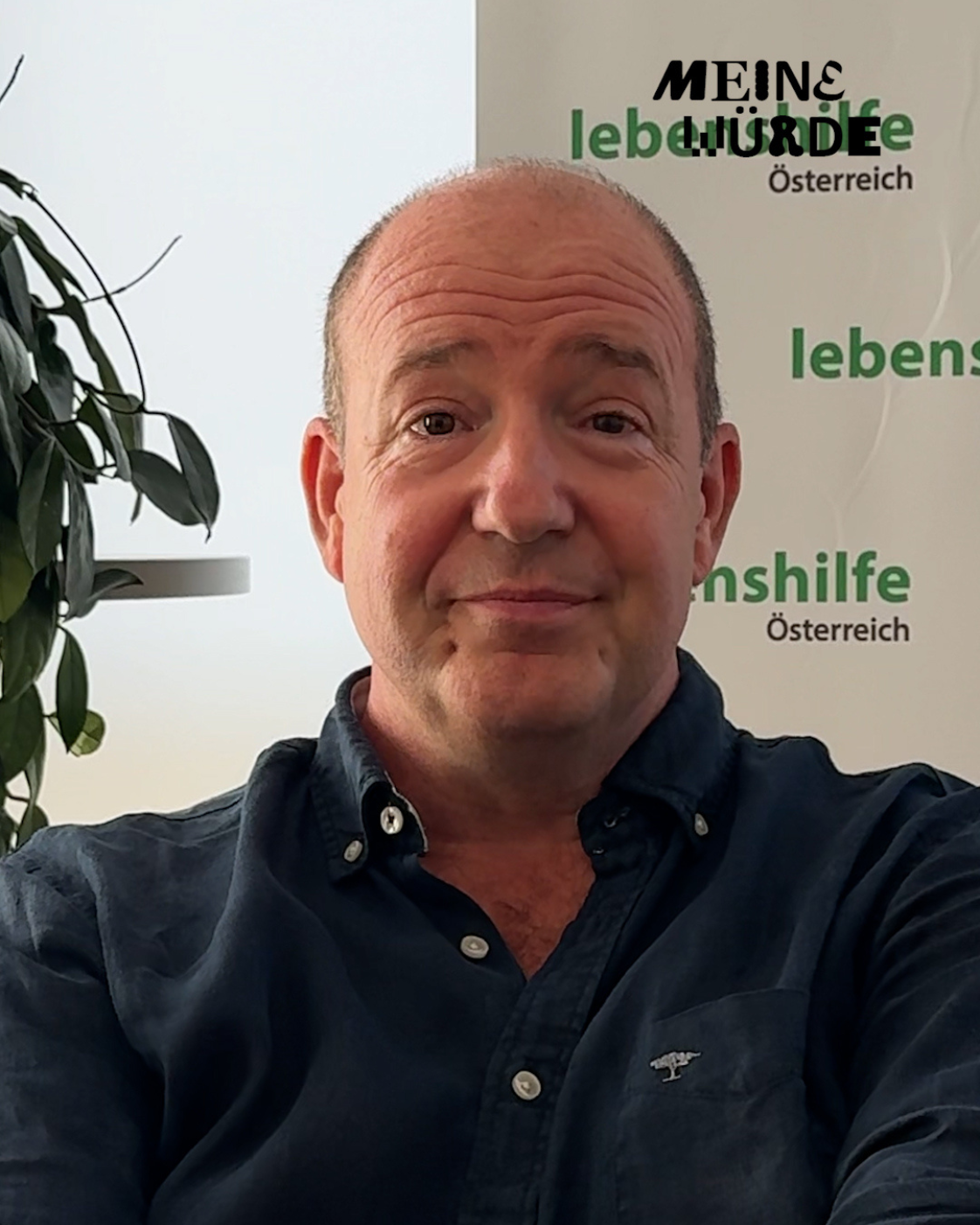 Mann mit dunklem Hemd, der frontal in die Kamera blickt. Im Hintergrund sind eine Zimmerpflanze und ein Aufsteller der Lebenshilfe Österreich zu sehen. Rechts oben befindet sich zusätzlich der Schriftzug „Meine Würde“.