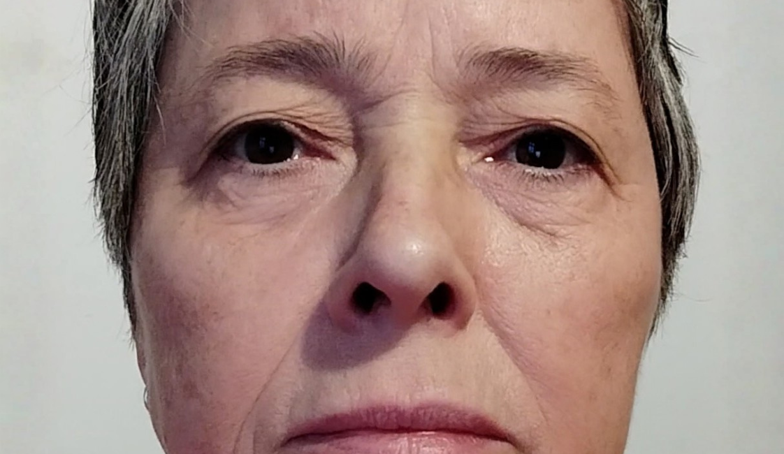 Nahaufnahme einer Frau mit kurzen grau-schwarzen Haaren und ernstem Gesichtsausdruck. Sie blickt frontal in die Kamera und trägt ein orangefarbenes Oberteil. Der Hintergrund ist hell und einfarbig.