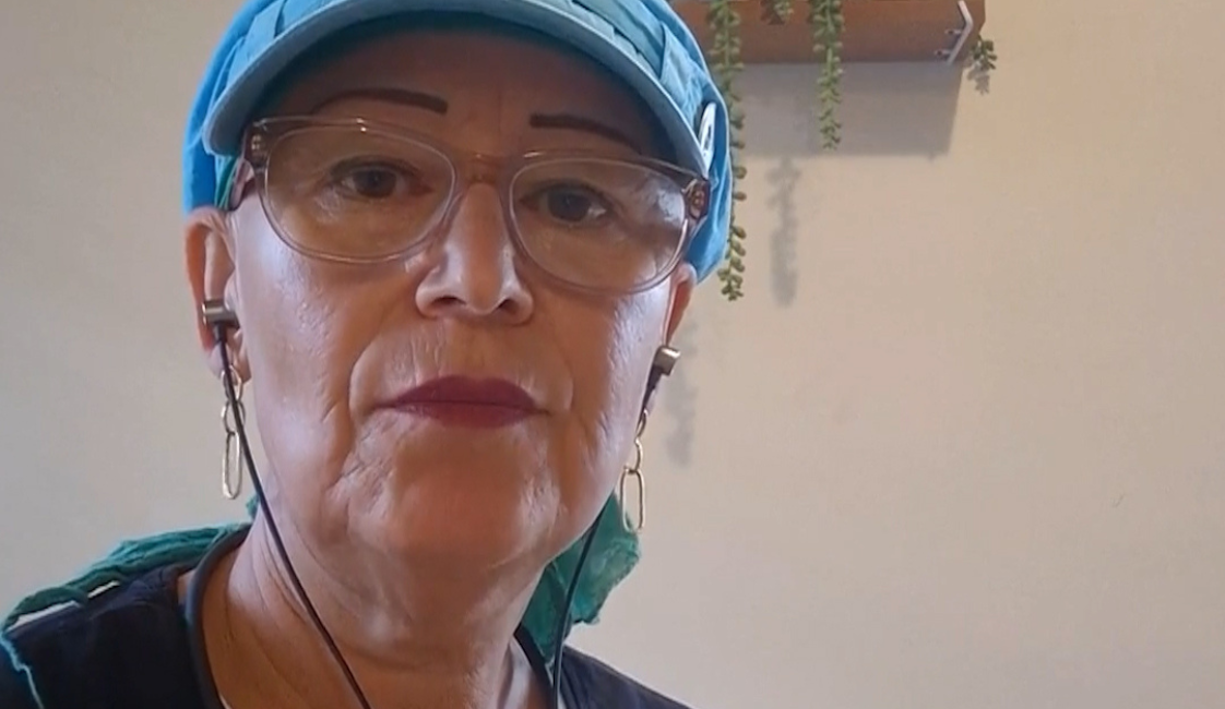 Frau mit blauer Kappe und Brille, die frontal in die Kamera blickt und Kopfhörer trägt. Im Hintergrund befinden sich zwei Wandregale mit Pflanzen und Dekoration. Rechts oben ist der Schriftzug „Meine Würde“ zu sehen.