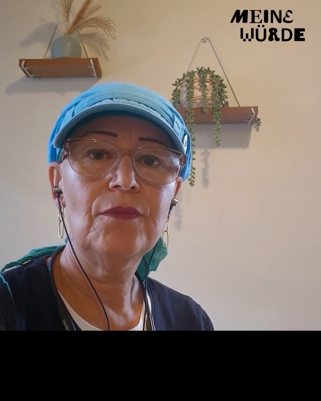 Frau mit blauer Kappe und Brille, die frontal in die Kamera blickt und Kopfhörer trägt. Im Hintergrund befinden sich zwei Wandregale mit Pflanzen und Dekoration. Rechts oben ist der Schriftzug „Meine Würde“ zu sehen.
