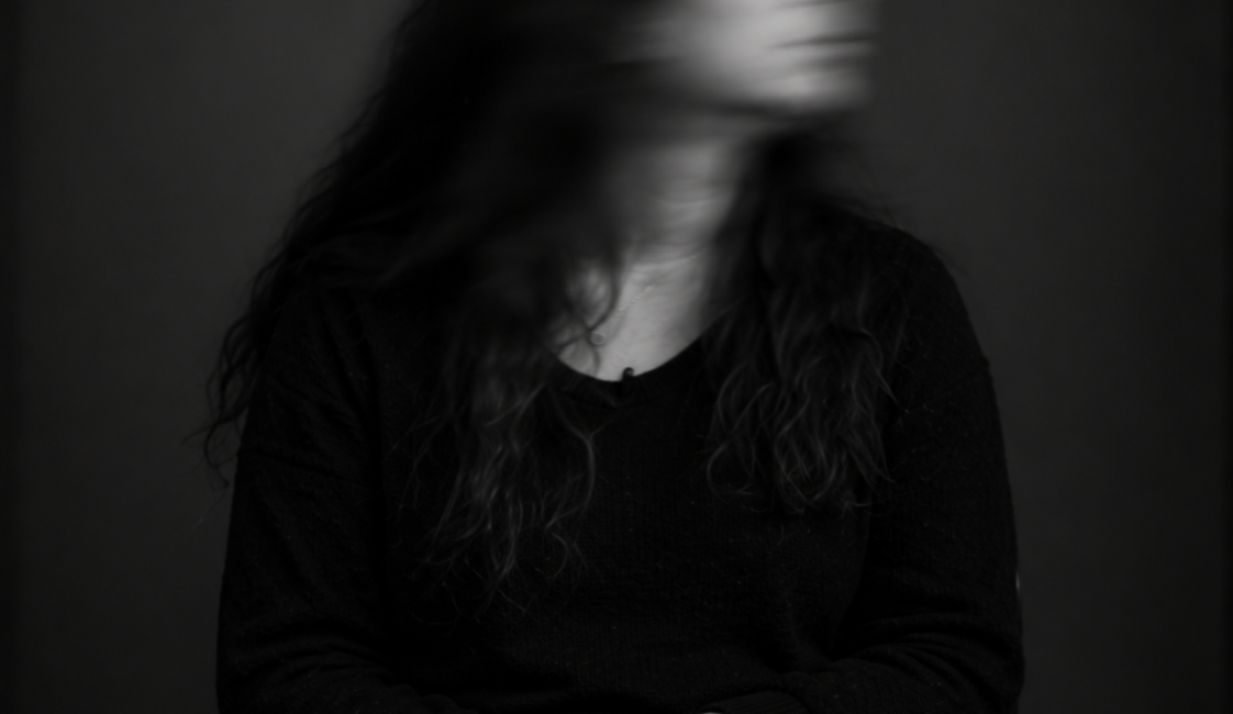 Sitzende Person mit langen dunklen Haaren und dunkler Kleidung. Das zur Seite gedrehte Gesicht ist durch Bewegungsunschärfe verschwommen.