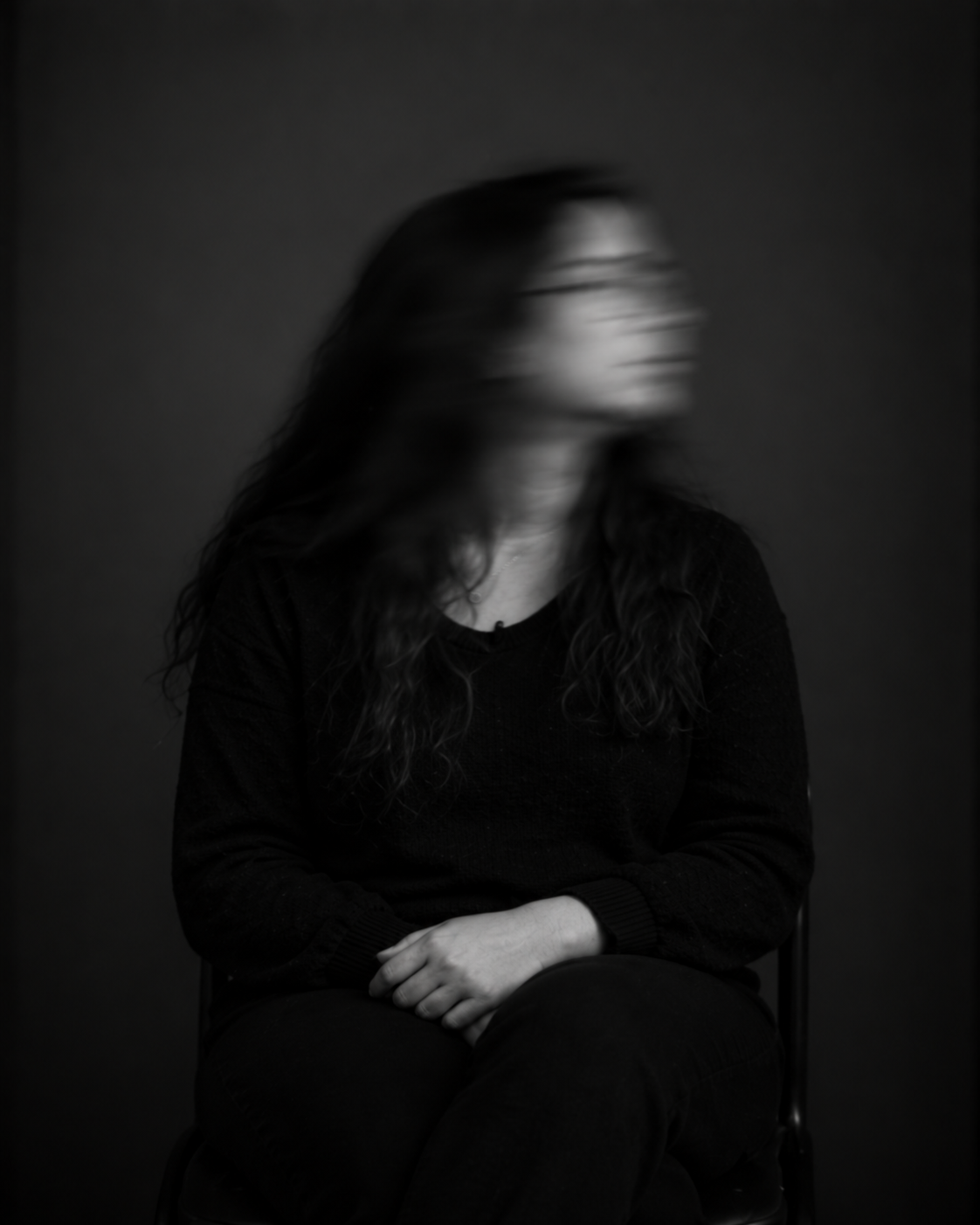 Sitzende Person mit langen dunklen Haaren und dunkler Kleidung. Das zur Seite gedrehte Gesicht ist durch Bewegungsunschärfe verschwommen.