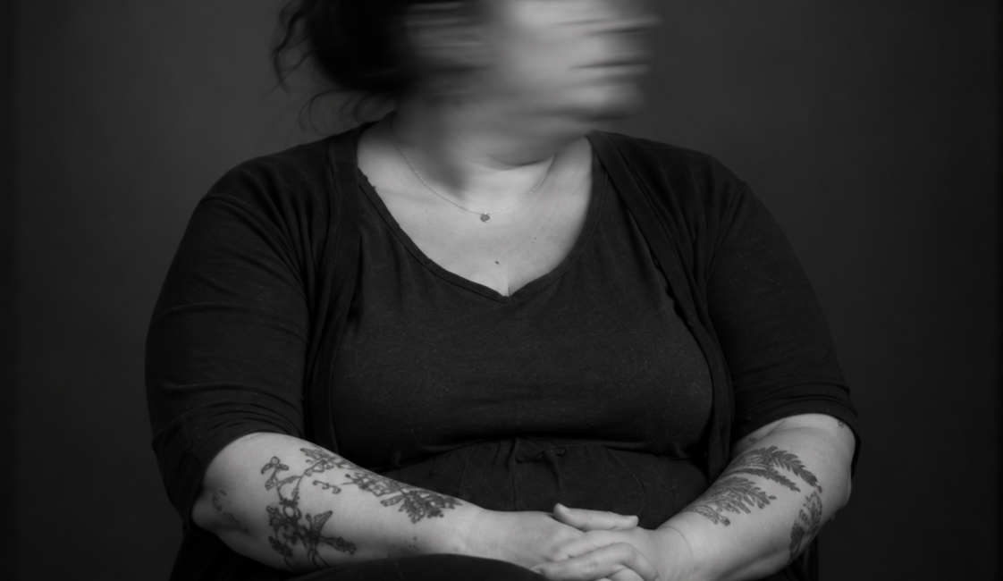 Sitzende Person in dunkler Kleidung mit verschränkten Händen im Schoß. Das zur Seite gedrehte Gesicht ist durch Bewegungsunschärfe verschwommen.
