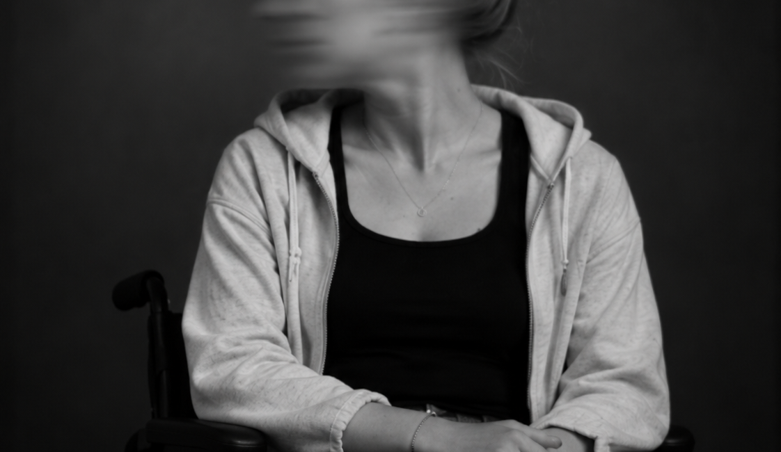 Sitzende Person im Rollstuhl mit hochgebundenen Haaren, heller Jacke und dunkler Kleidung. Das zur Seite gedrehte Gesicht ist durch Bewegungsunschärfe verschwomme