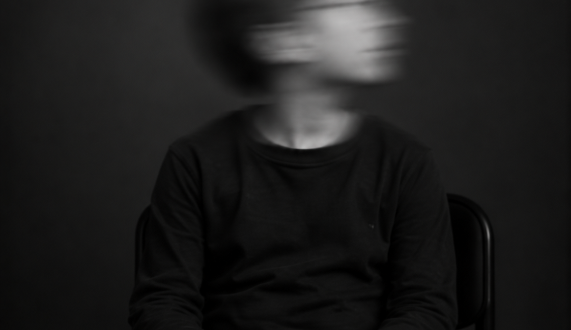 Sitzende Person mit kurzen dunklen Haaren, dunklem Pullover und Jeans. Das zur Seite gedrehte Gesicht ist durch Bewegungsunschärfe verschwommen.