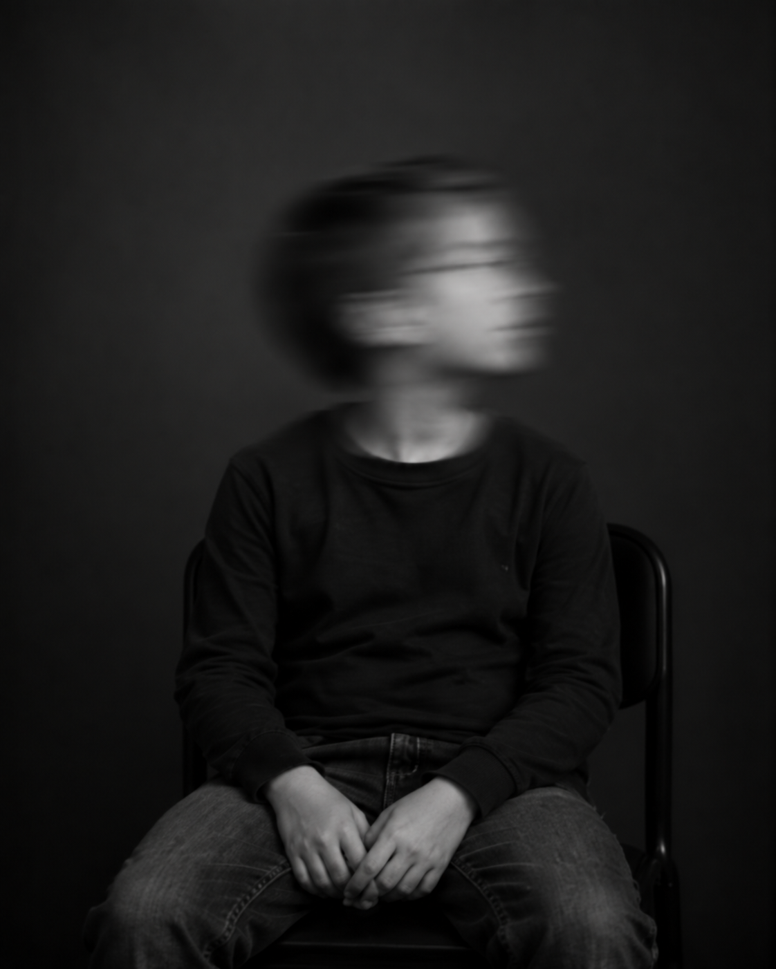 Sitzende Person mit kurzen dunklen Haaren, dunklem Pullover und Jeans. Das zur Seite gedrehte Gesicht ist durch Bewegungsunschärfe verschwommen.