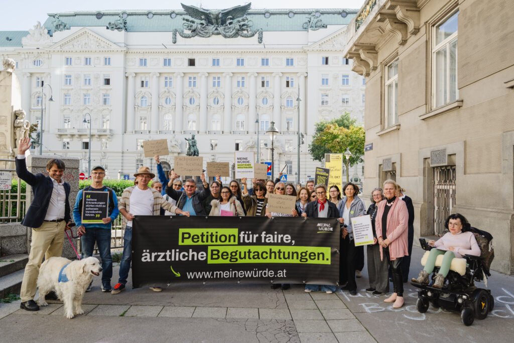 Gruppe von Demonstrant:innen bei einer Kundgebung für faire ärztliche Begutachtungen. Sie halten ein großes Banner mit der Aufschrift „Petition für faire, ärztliche Begutachtungen“ sowie verschiedene Protestschilder. Rechts sitzt eine Person im Rollstuhl, links steht ein Assistenzhund.