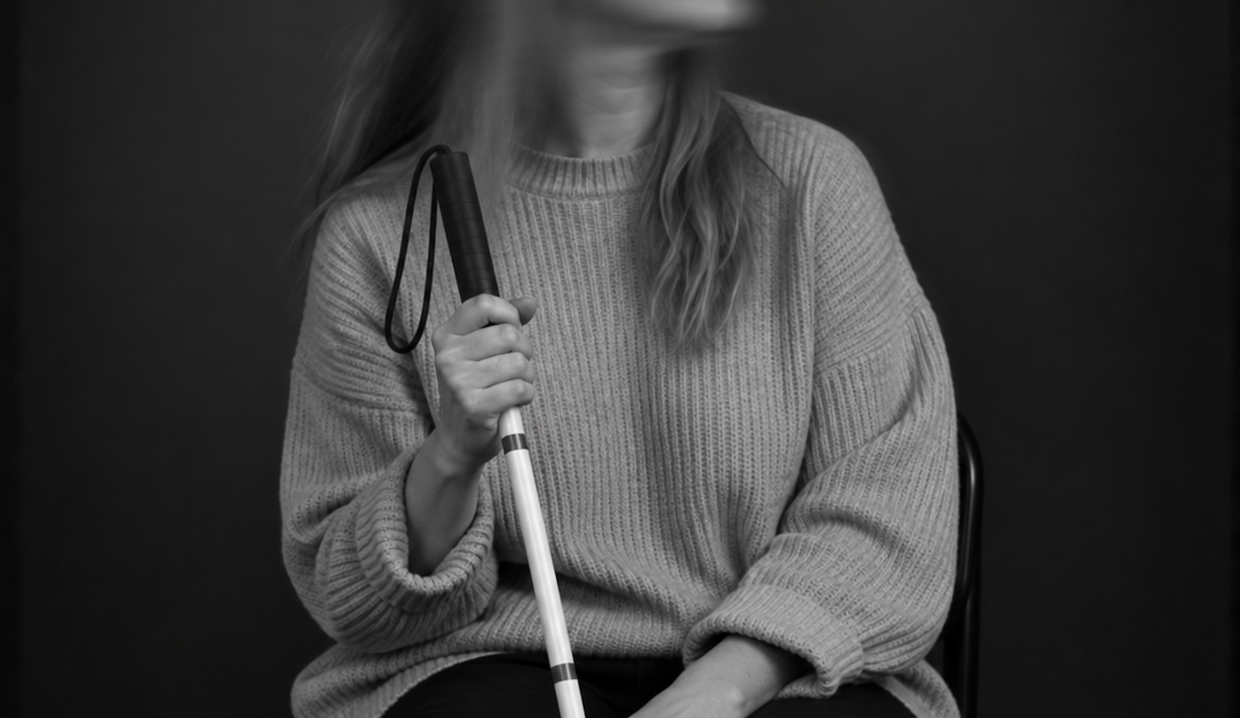 Sitzende Person mit langen Haaren, Sonnenbrille und heller Jacke. Sie hält einen weißen Langstock in der Hand und blickt zur Seite.