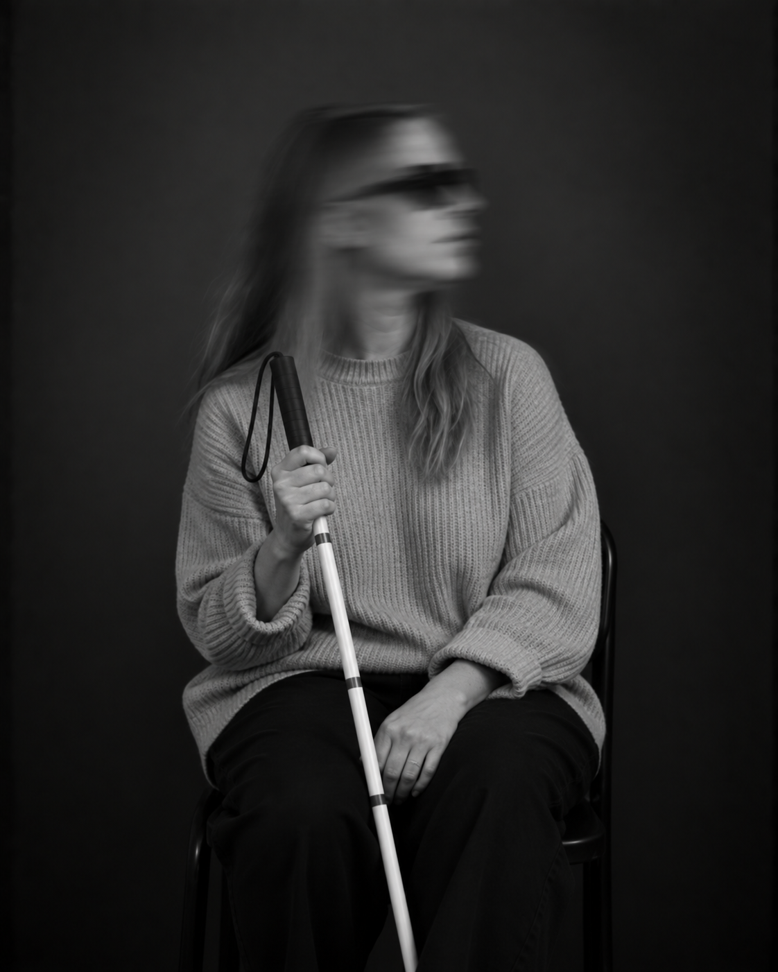Sitzende Person mit langen Haaren, Sonnenbrille und heller Jacke. Sie hält einen weißen Langstock in der Hand und blickt zur Seite.