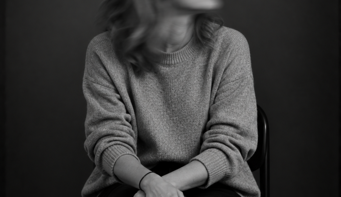 Sitzende Person mit langen Haaren, heller Jacke und dunkler Kleidung. Das zur Seite gedrehte Gesicht ist durch Bewegungsunschärfe verschwommen.