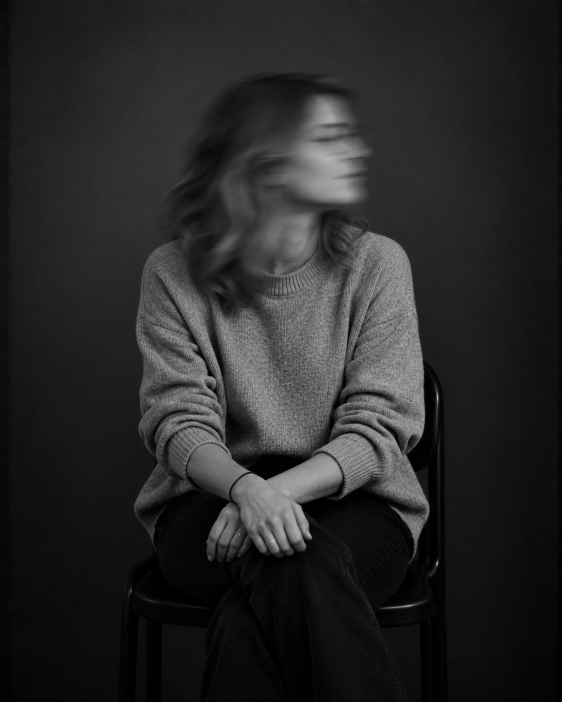 Sitzende Person mit langen Haaren, heller Jacke und dunkler Kleidung. Das zur Seite gedrehte Gesicht ist durch Bewegungsunschärfe verschwommen.
