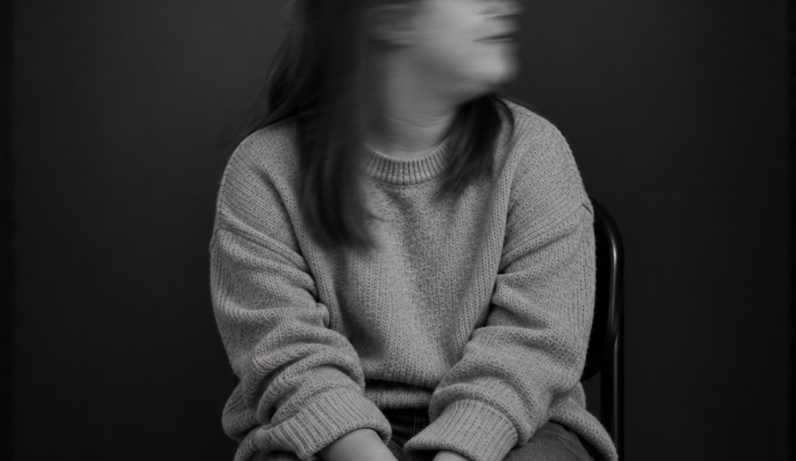 Sitzende Person mit langen Haaren und heller Kleidung, die beide Hände auf den Knien hält. Das zur Seite gedrehte Gesicht ist durch Bewegungsunschärfe verschwommen.