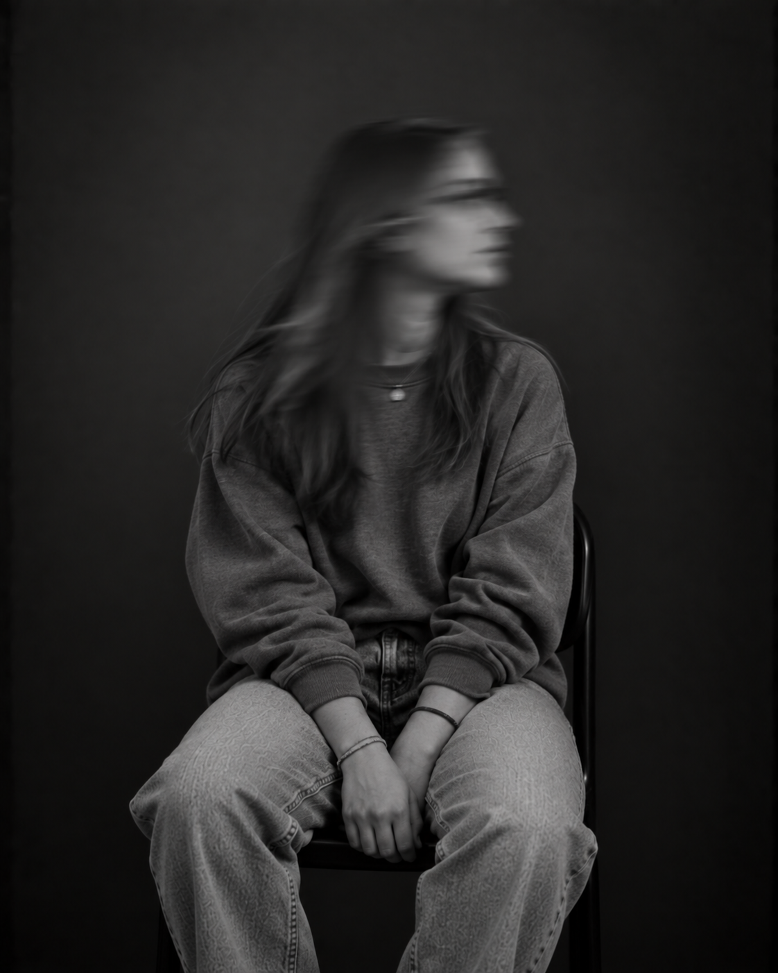 Sitzende Person mit langen Haaren, dunkler Jacke und heller Hose, die beide Hände zwischen den Knien hält. Das zur Seite gedrehte Gesicht ist durch Bewegungsunschärfe verschwommen.
