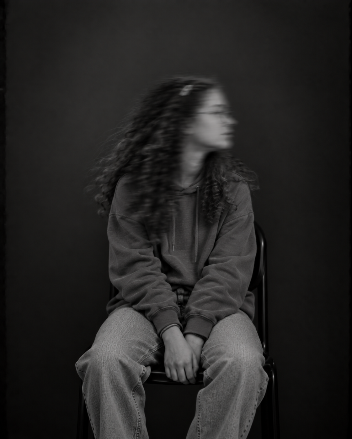 Sitzende Person mit langen dunklen Haaren, dunklem Pullover und heller Hose. Das zur Seite gedrehte Gesicht ist durch Bewegungsunschärfe verschwommen.