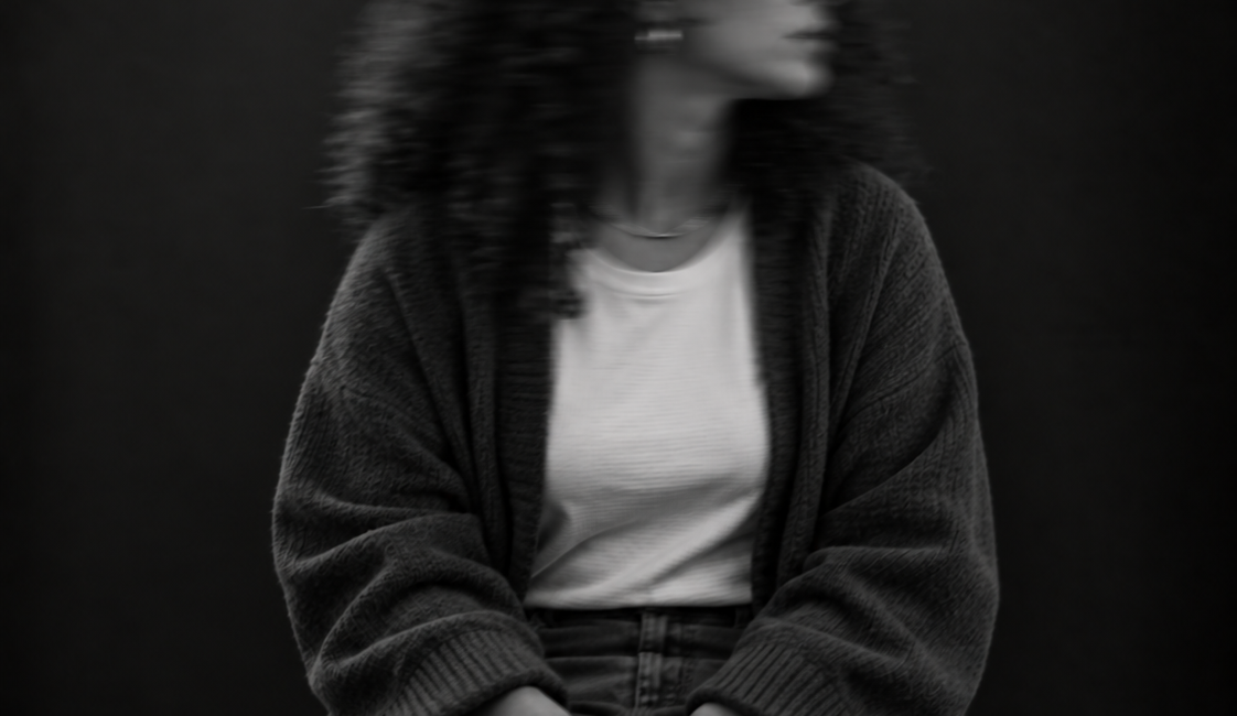 Sitzende Person mit langen lockigen Haaren, heller Bluse und dunkler Jacke. Das zur Seite gedrehte Gesicht ist durch Bewegungsunschärfe verschwommen.