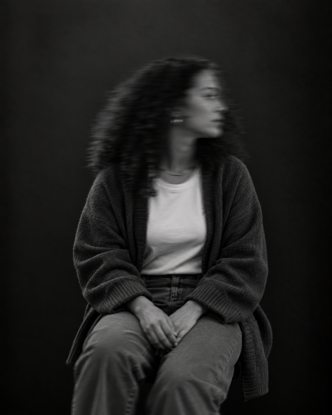 Sitzende Person mit langen lockigen Haaren, heller Bluse und dunkler Jacke. Das zur Seite gedrehte Gesicht ist durch Bewegungsunschärfe verschwommen.