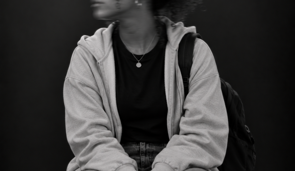 Sitzende Person mit hochgebundenen Haaren, heller Jacke und dunkler Kleidung. Das zur Seite gedrehte Gesicht ist durch Bewegungsunschärfe verschwommen.