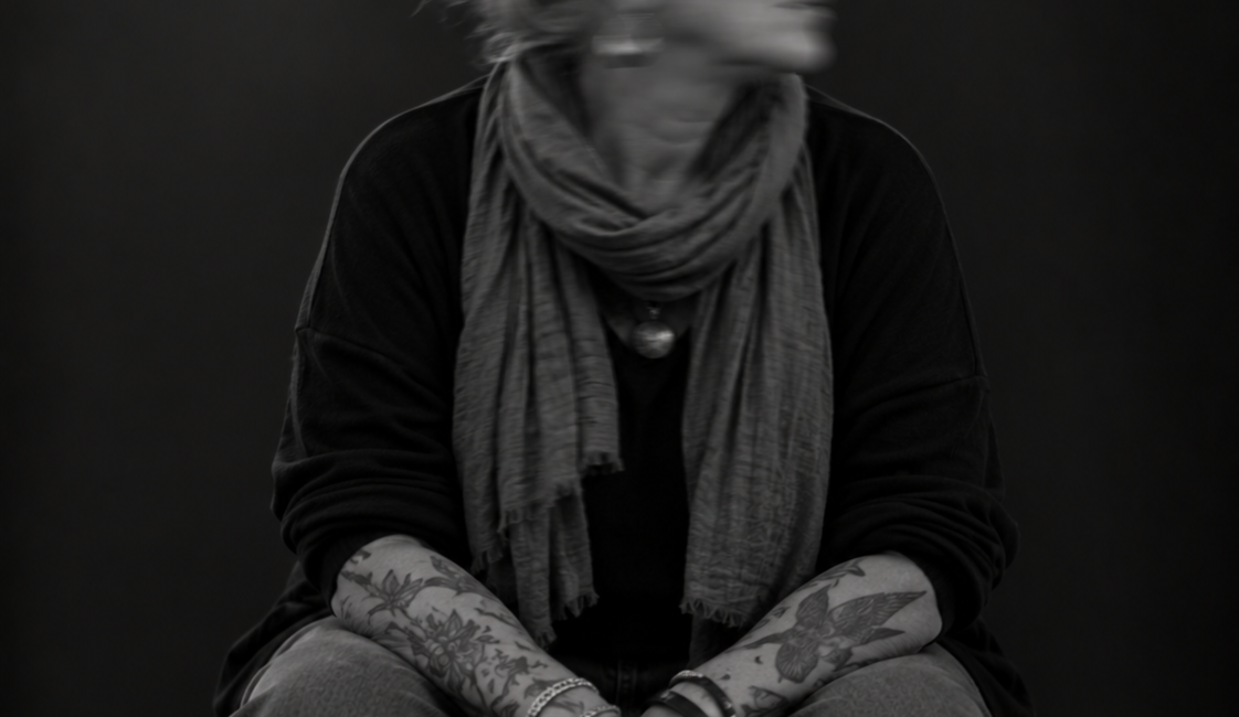 Sitzende Person mit Kopftuch, Schal und dunkler Kleidung, die beide Hände zwischen den Knien hält. Das nach unten gedrehte Gesicht ist durch Bewegungsunschärfe verschwommen.