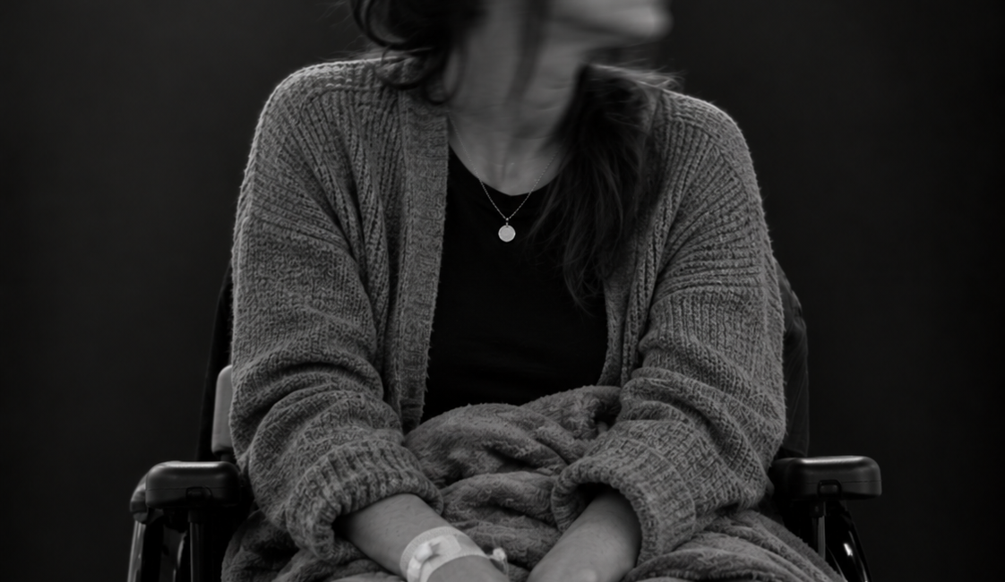 Sitzende Person im Rollstuhl mit schulterlangen Haaren und heller Jacke. Das zur Seite gedrehte Gesicht ist durch Bewegungsunschärfe verschwommen.