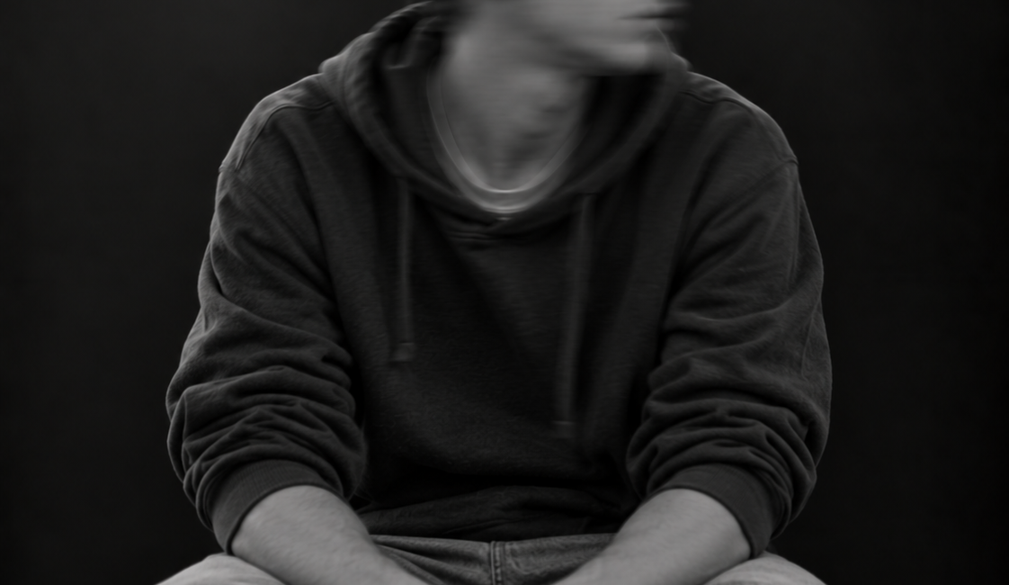 Sitzende Person mit kurzen lockigen Haaren, dunklem Pullover und heller Hose. Das zur Seite geneigte Gesicht ist durch Bewegungsunschärfe verschwommen.