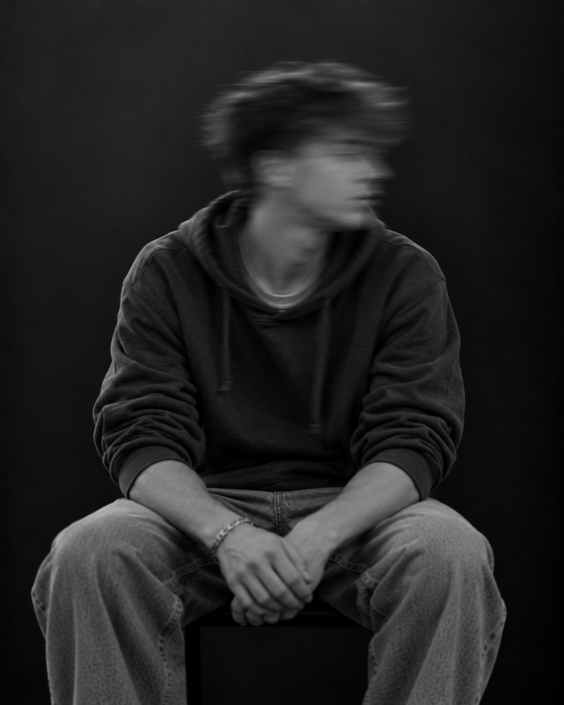 Sitzende Person mit kurzen lockigen Haaren, dunklem Pullover und heller Hose. Das zur Seite geneigte Gesicht ist durch Bewegungsunschärfe verschwommen.