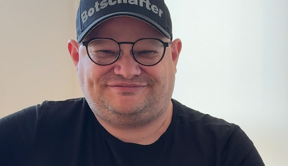 Sitzender Mann mit Brille, schwarzem T-Shirt und schwarzer Kappe mit der Aufschrift „Botschafter“. Rechts oben steht „Meine Würde“.
