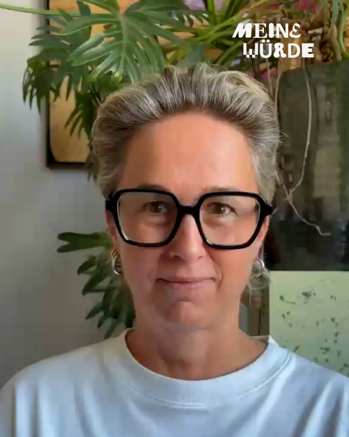 Person mit kurzen grauen Haaren und großer schwarzer Brille vor grünen Zimmerpflanzen. Rechts oben steht „Meine Würde“.