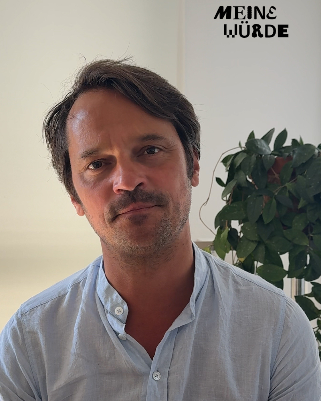 Person mit kurzen dunklen Haaren und hellem Hemd, die frontal in die Kamera blickt. Im Hintergrund steht eine grüne Zimmerpflanze. Rechts oben steht „Meine Würde“.