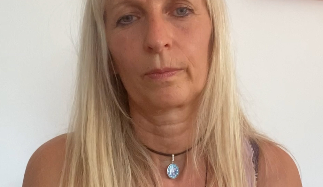 Person mit langen blonden Haaren und lilafarbenem Oberteil, die frontal in die Kamera blickt. Sie trägt zwei Halsketten mit Anhängern. Im Hintergrund ist eine weiße Wand mit einem farbigen Bild zu sehen.