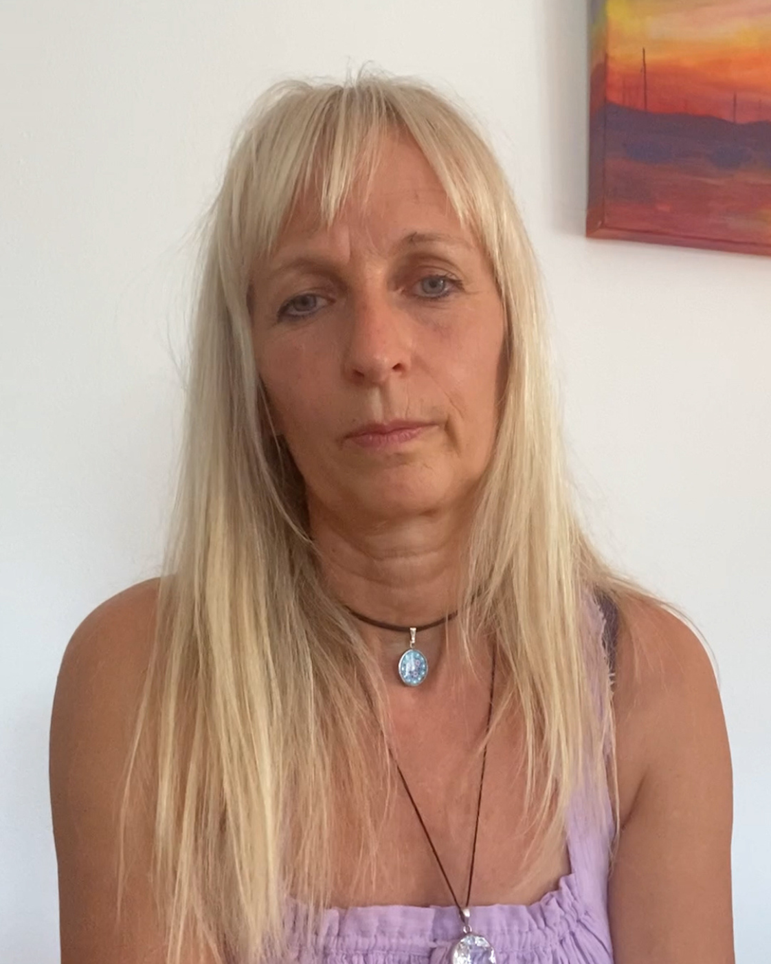 Person mit langen blonden Haaren und lilafarbenem Oberteil, die frontal in die Kamera blickt. Sie trägt zwei Halsketten mit Anhängern. Im Hintergrund ist eine weiße Wand mit einem farbigen Bild zu sehen.