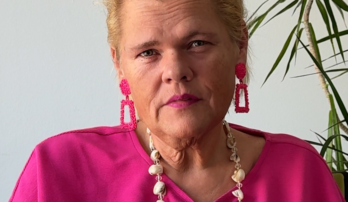 Person mit zurückgebundenen blonden Haaren und pinkem Oberteil, die frontal in die Kamera blickt. Sie trägt auffällige pinke Ohrringe und eine helle Halskette; eine Sonnenbrille hängt am Oberteil. Im Hintergrund ist eine Zimmerpflanze zu sehen.