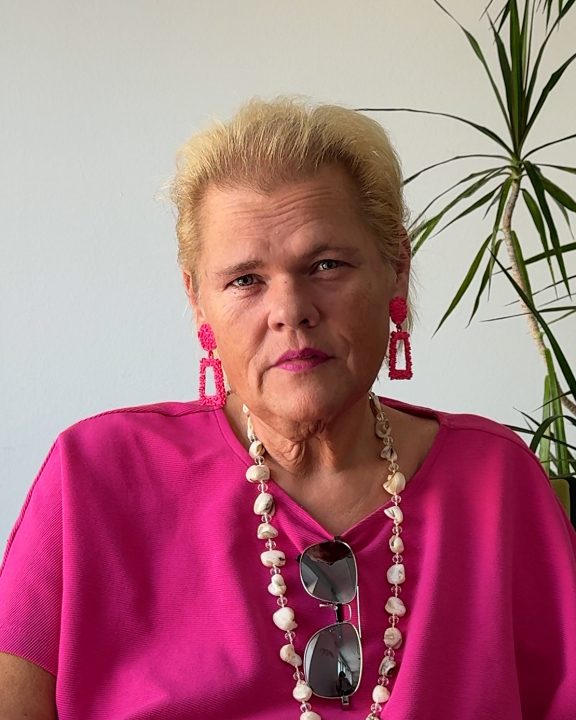 Person mit zurückgebundenen blonden Haaren und pinkem Oberteil, die frontal in die Kamera blickt. Sie trägt auffällige pinke Ohrringe und eine helle Halskette; eine Sonnenbrille hängt am Oberteil. Im Hintergrund ist eine Zimmerpflanze zu sehen.