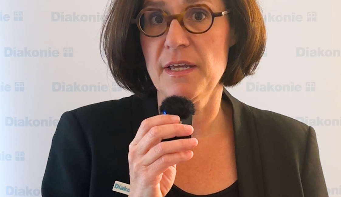 Person mit kurzem dunklem Bob und Brille, die ein kleines Mikrofon in der Hand hält und in die Kamera spricht. Sie trägt einen schwarzen Blazer. Im Hintergrund ist mehrfach der Schriftzug „Diakonie“ zu sehen.