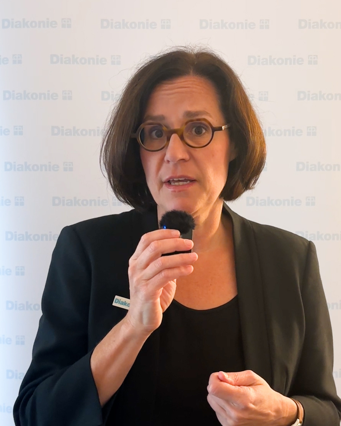 Person mit kurzem dunklem Bob und Brille, die ein kleines Mikrofon in der Hand hält und in die Kamera spricht. Sie trägt einen schwarzen Blazer. Im Hintergrund ist mehrfach der Schriftzug „Diakonie“ zu sehen.