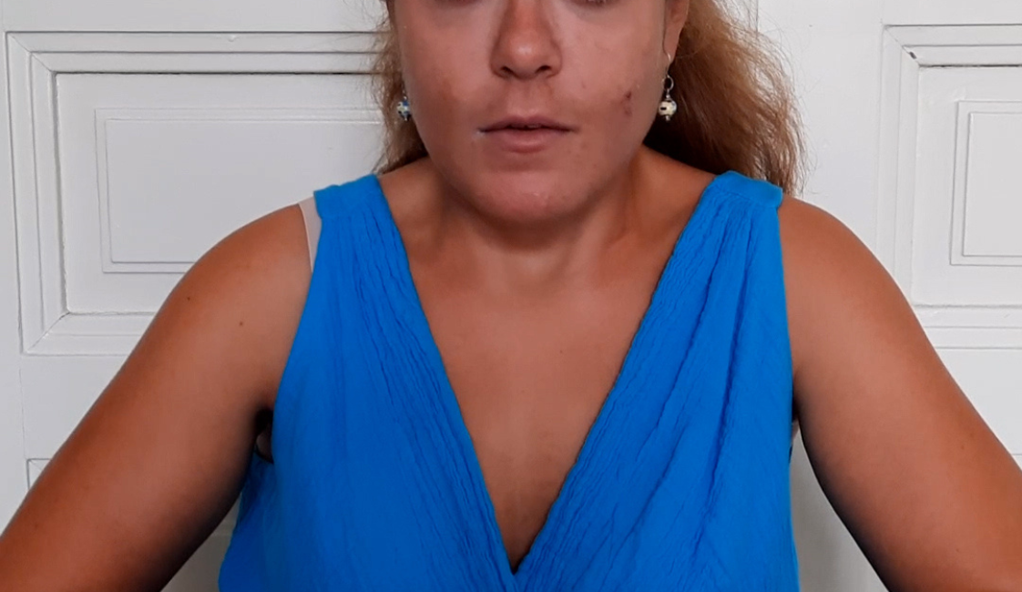 Person mit zurückgebundenen braunen Haaren und leuchtend blauem Kleid, die frontal in die Kamera blickt. Sie sitzt vor einer weißen Flügeltür und hält die Hände vor dem Körper. Rechts oben steht „Meine Würde“.