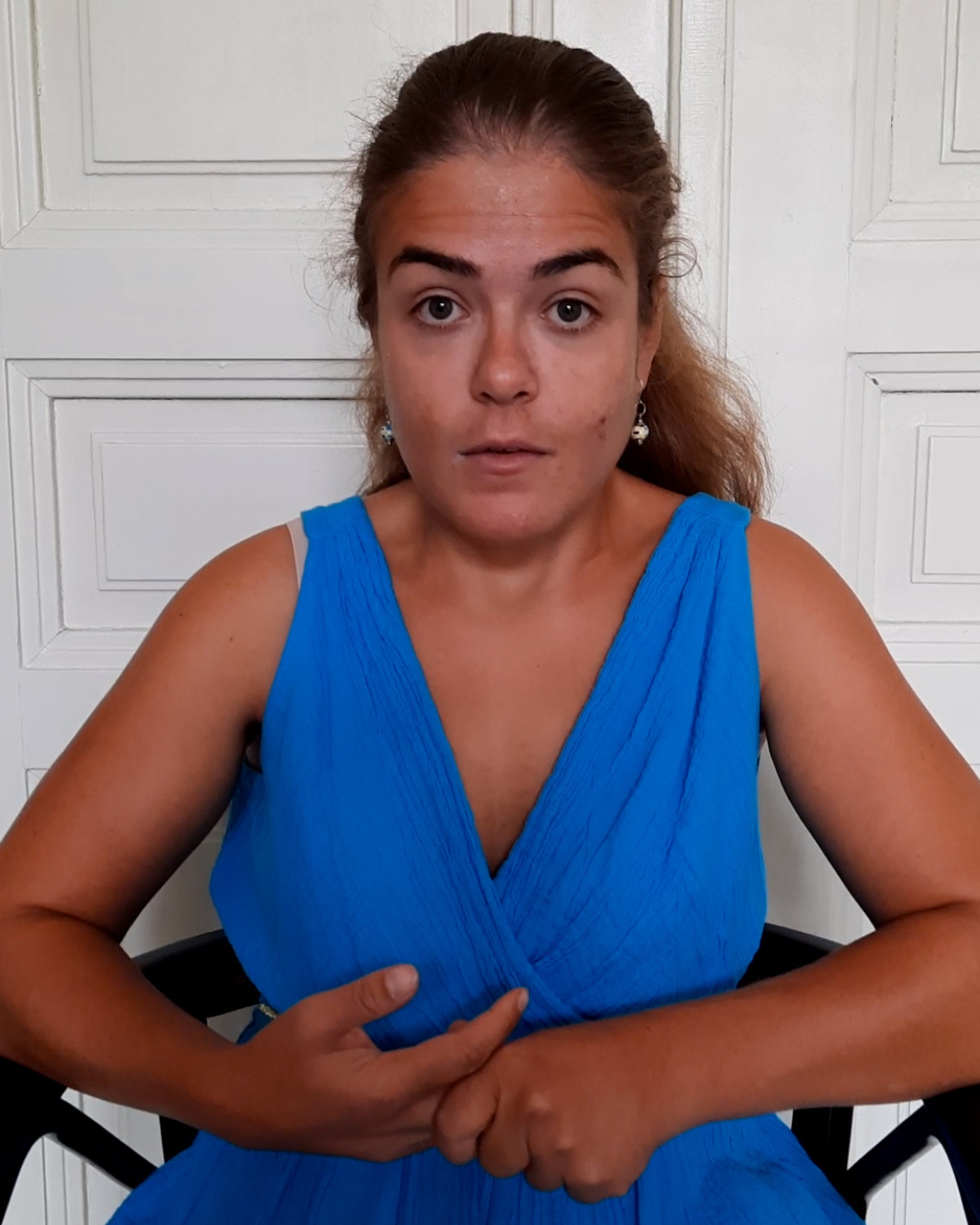 Person mit zurückgebundenen braunen Haaren und leuchtend blauem Kleid, die frontal in die Kamera blickt. Sie sitzt vor einer weißen Flügeltür und hält die Hände vor dem Körper. Rechts oben steht „Meine Würde“.