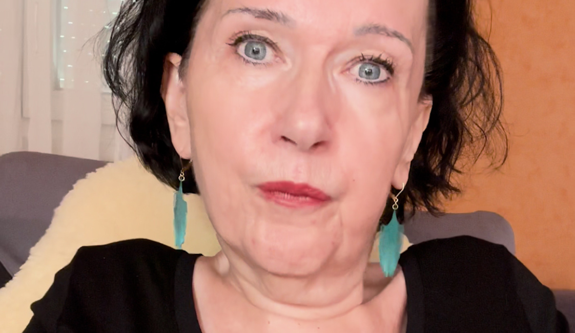 Frau mit kurzen schwarzen Haaren, türkisfarbenen Ohrringen und einem schwarzen Oberteil mit Blumenmuster. Sie sitzt in einem Sessel und blickt frontal in die Kamera. Rechts oben befindet sich der weiße Schriftzug „Meine Würde“.