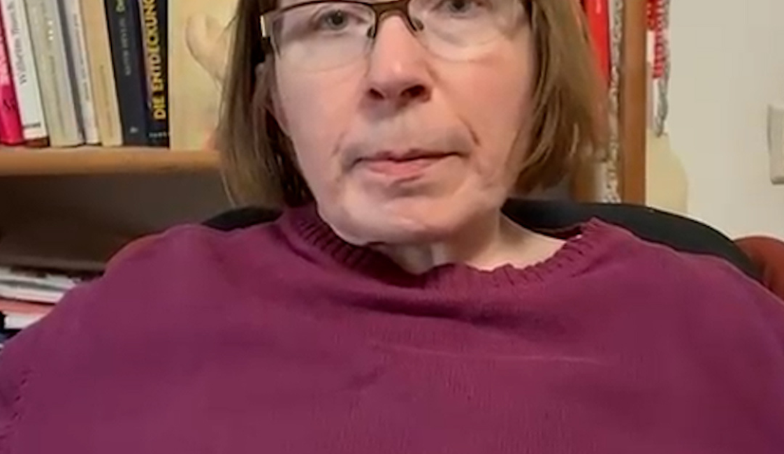 Eine Frau mit Brille und kurzem braunem Haar sitzt vor einem Bücherregal. Sie trägt einen dunkelroten Pullover und blickt in die Kamera.
