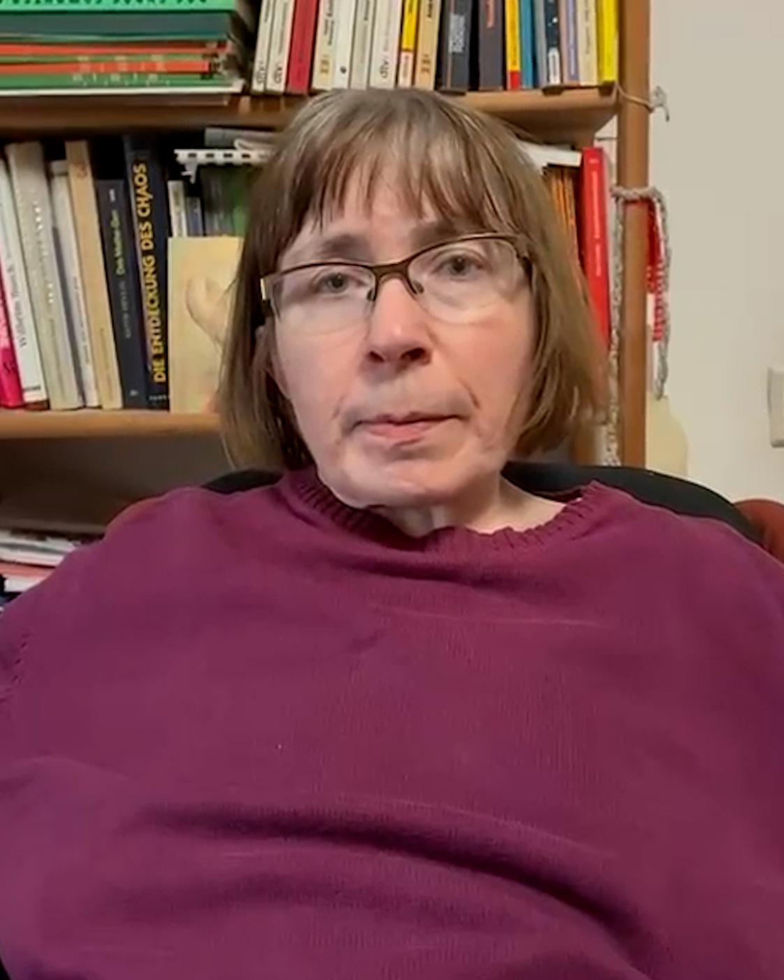 Eine Frau mit Brille und kurzem braunem Haar sitzt vor einem Bücherregal. Sie trägt einen dunkelroten Pullover und blickt in die Kamera.