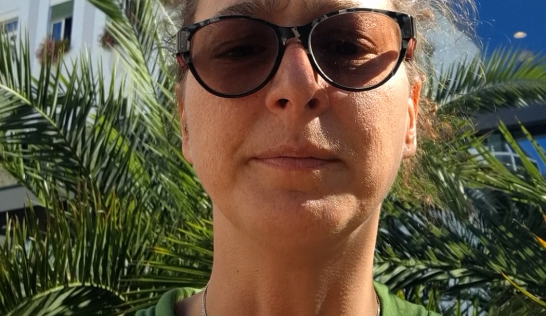 Frau mit dunkler Sonnenbrille und grünem Oberteil, die draußen frontal in die Kamera blickt. Hinter ihr sind Palmen, Gebäude und ein blauer Himmel zu sehen. Rechts oben befindet sich der weiße Schriftzug „Meine Würde“.