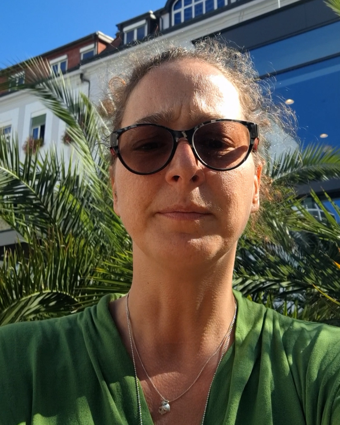 Frau mit dunkler Sonnenbrille und grünem Oberteil, die draußen frontal in die Kamera blickt. Hinter ihr sind Palmen, Gebäude und ein blauer Himmel zu sehen. Rechts oben befindet sich der weiße Schriftzug „Meine Würde“.