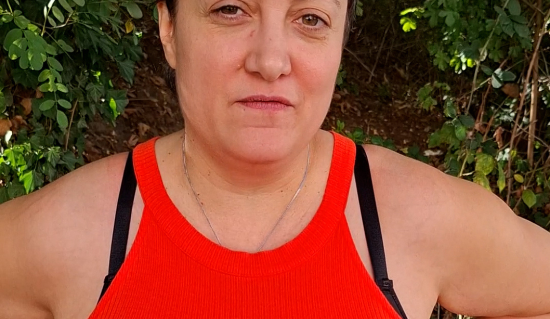 Frau mit kurzen dunklen Haaren und orangefarbenem Oberteil, die draußen mit den Händen in den Hüften frontal in die Kamera blickt. Im Hintergrund sind dichte grüne Pflanzen zu sehen. Rechts oben befindet sich der weiße Schriftzug „Meine Würde“.