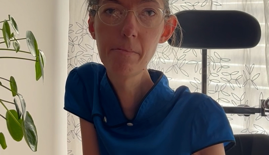 Eine Frau sitzt in einem Rollstuhl vor einem hellen Fenster mit einem gemusterten Vorhang. Sie trägt eine blaue Bluse und eine helle Brille. Links im Bild ist eine Zimmerpflanze zu sehen.