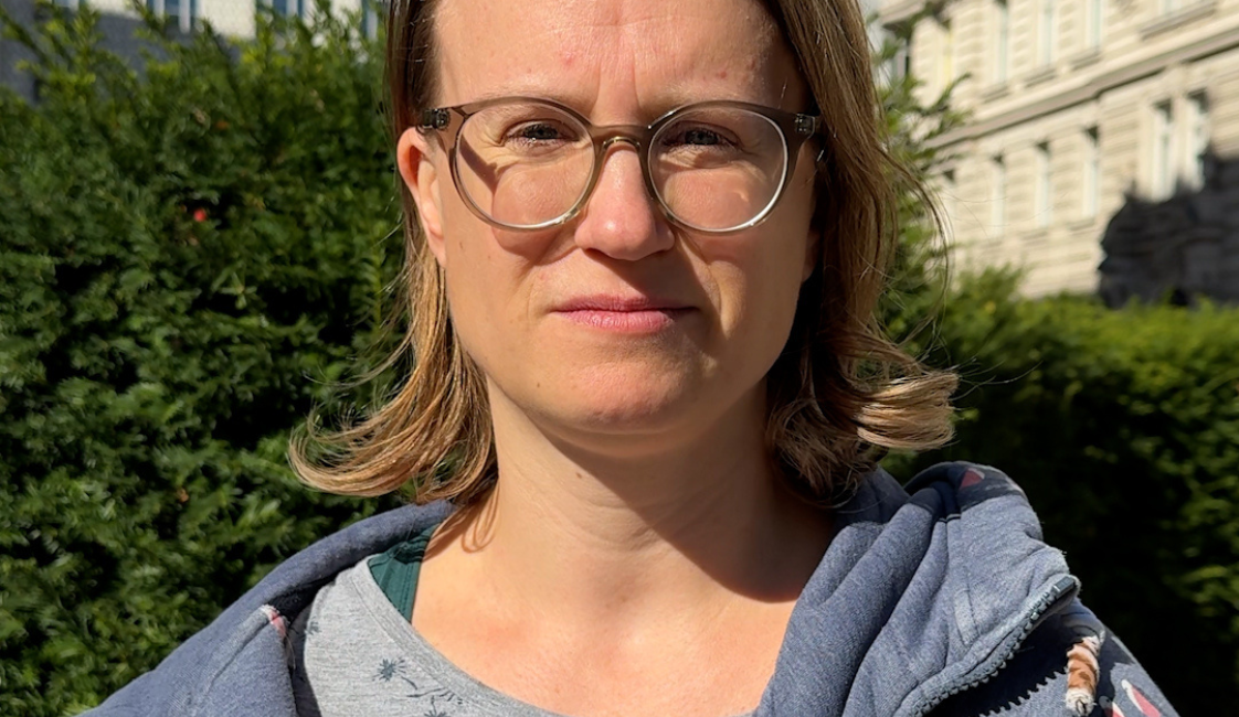 Eine Frau mit schulterlangen braunen Haaren und Brille steht im Freien und blickt in die Kamera. Sie trägt eine blaue Kapuzenjacke mit Vogelmuster. Im Hintergrund sind grüne Sträucher und das Gebäude der Österreichischen Postsparkasse zu sehen.