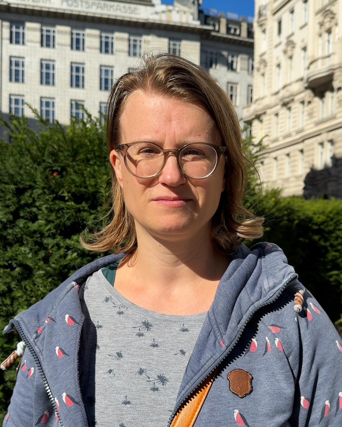 Eine Frau mit schulterlangen braunen Haaren und Brille steht im Freien und blickt in die Kamera. Sie trägt eine blaue Kapuzenjacke mit Vogelmuster. Im Hintergrund sind grüne Sträucher und das Gebäude der Österreichischen Postsparkasse zu sehen.