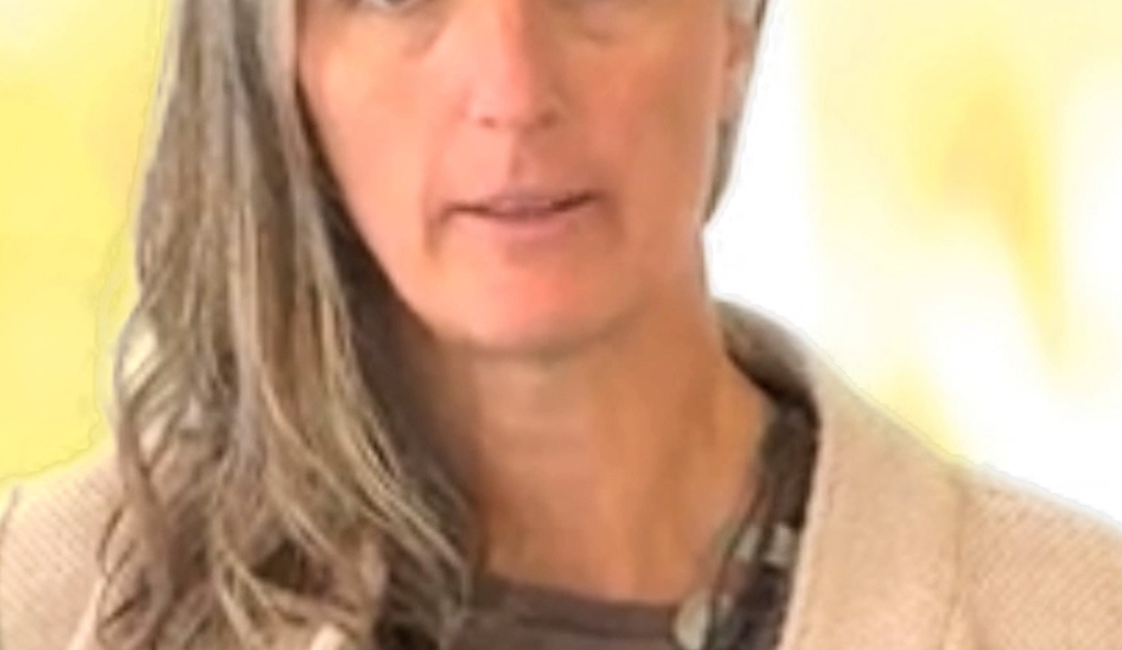 Eine Frau mit langen grauen Haaren blickt in die Kamera. Sie trägt ein graubraunes Oberteil, eine beige Strickjacke und eine Halskette. An ihrer Kleidung ist ein kleines Mikrofon befestigt. Im Hintergrund ist eine helle Wand zu sehen.